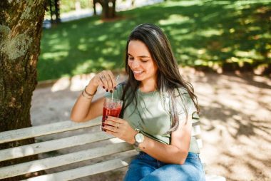 Smiling woman sitting on a bench enjoying a cold beverage in a sunny park.